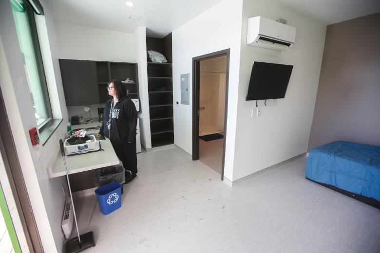 A woman stands in a white room, in front of a sink and window with a bed behind her