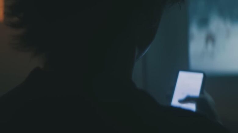 A person, pictured from behind, is shown holding a smart phone in a darkened room.  