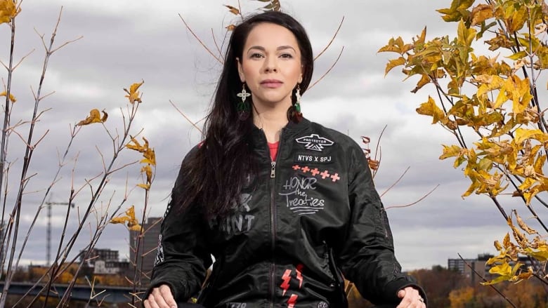 An Indigenous woman wearing a black zip-up and leggings sits cross legged, with her hands on her knees, outside. 