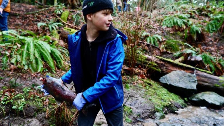 A boy in a blue jacket gets ready to throw a salmon into a forested creek.