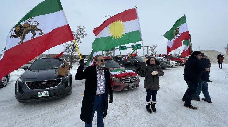 Four people are lined up in front of several vehicles. Each of them are waving Iranian flags.