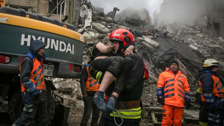 An emergency worker carries a woman while several other rescue workers are seen in the background, in front of a very large pile of rubble.