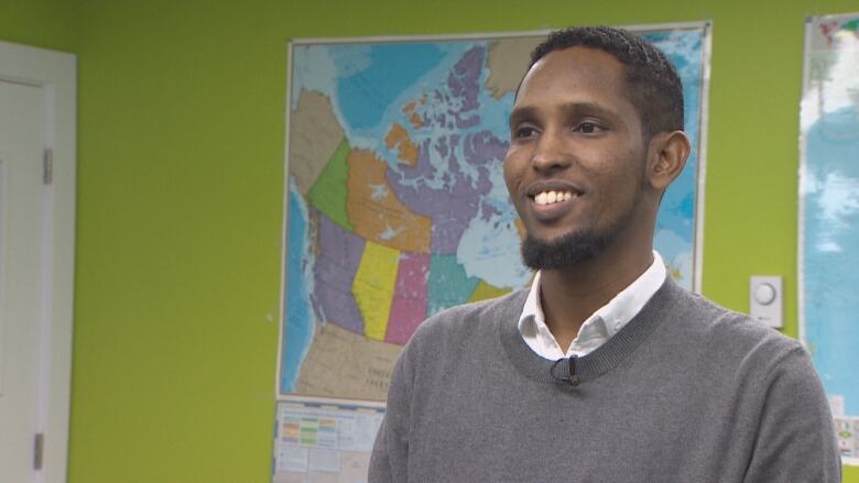 A man in a grey sweater with a white shirt is smiling as he gives an interview with a map of the world on a wall behind him.