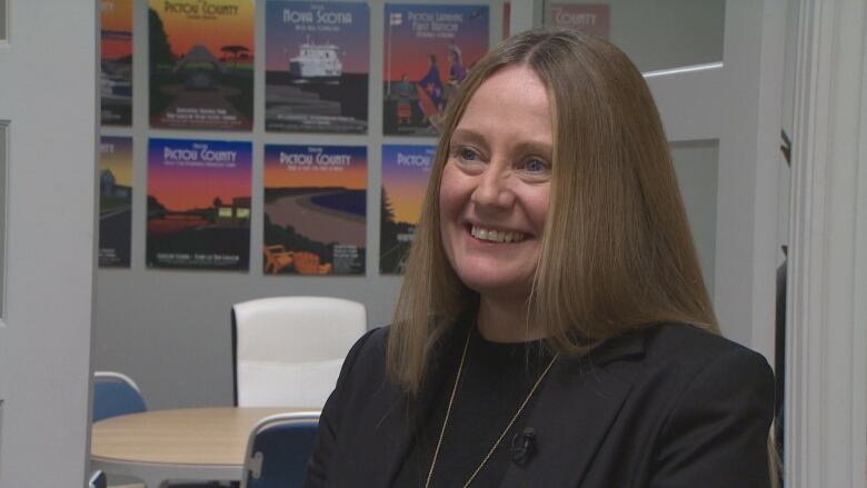 A woman is shown smiling with posters of Pictou County on a wall behind her.