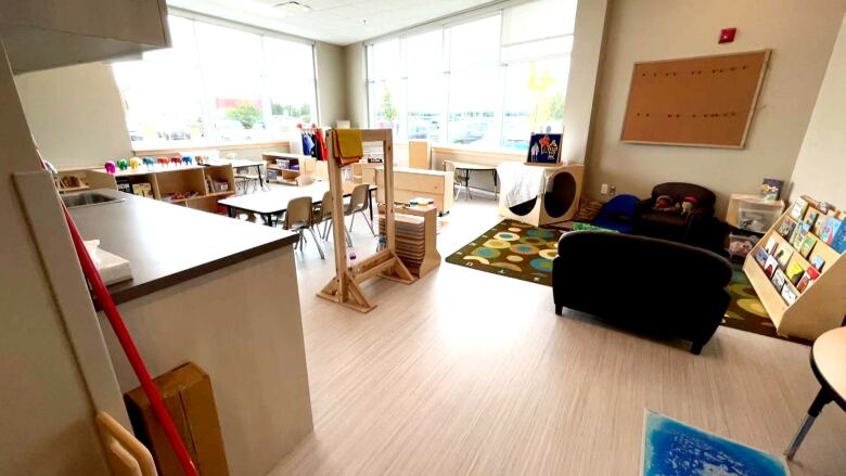 A child care room has tables, chairs, couches, books and a colourful rug with toys spread around. 