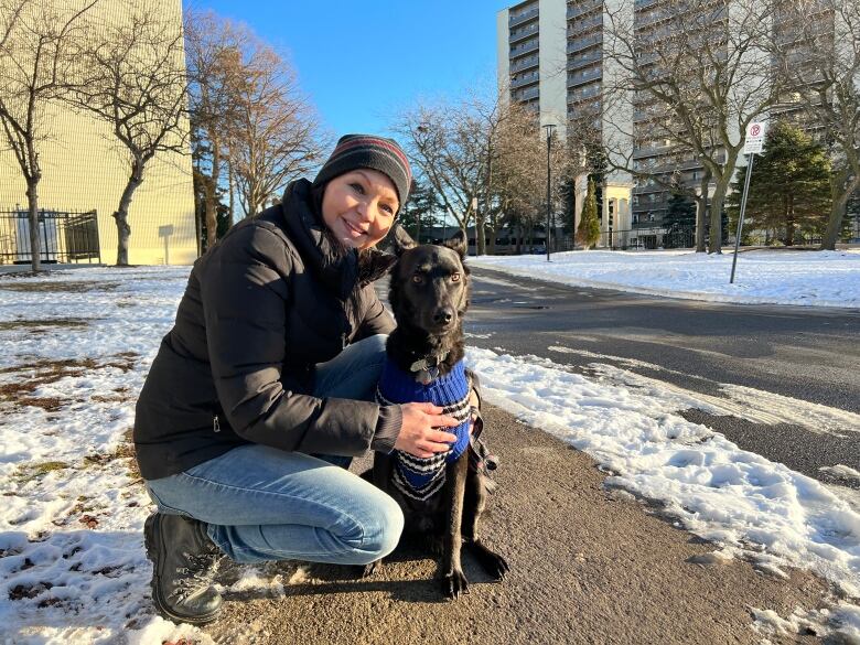 A woman kneeling and smiling at the camera, with a black dog sitting beside her. 