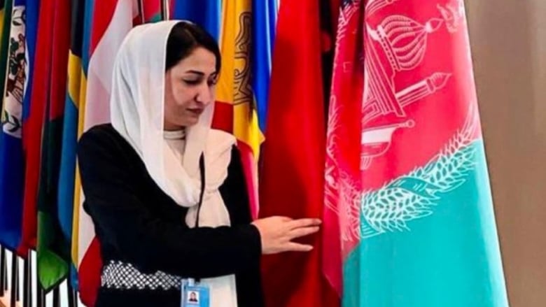 A woman in a black dress stands next to a row of national flags, gently touching the Afghanistan one.