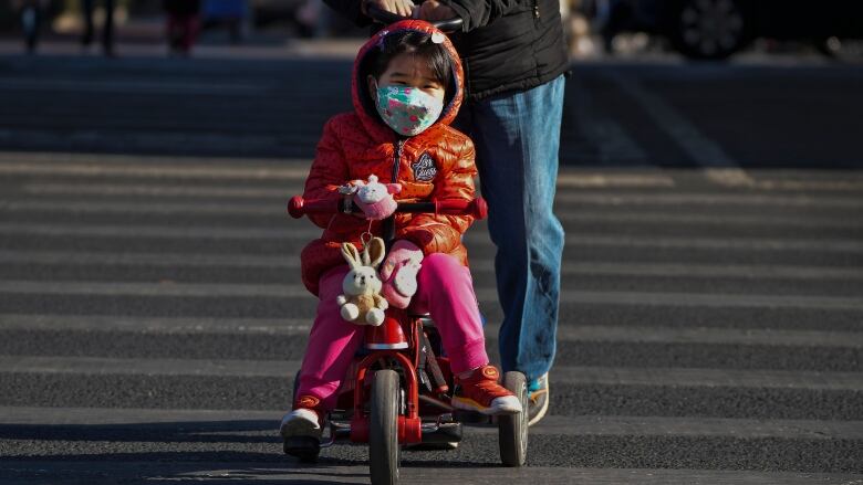 A child wearing a red jacket and face mask rides a tricycle towards the camera.