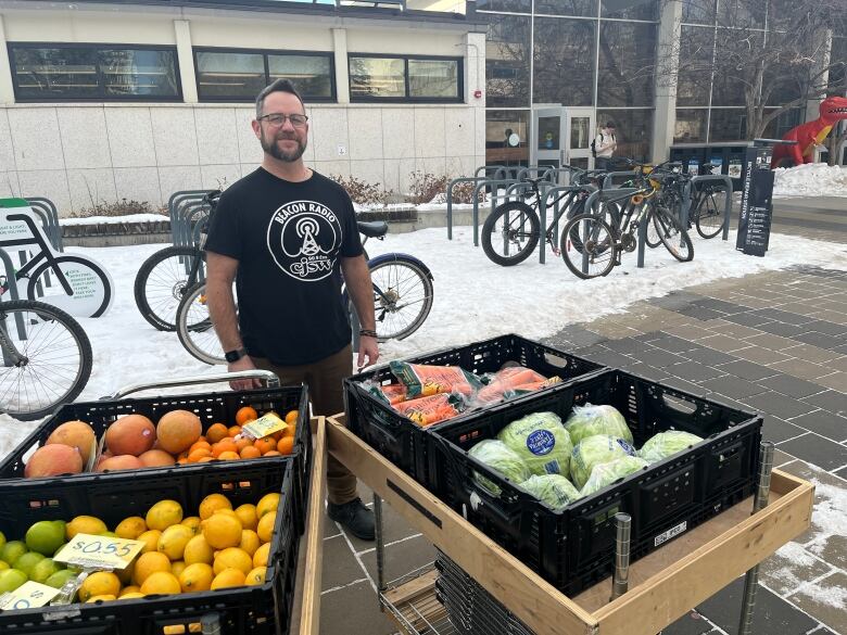 Robillard stands in a black t-shirt emblazoned with the CJSW logo, beside boxes of fruits and vegetables on carts. He is outside, with snow and locked bikes in the background, along with a red dinosaur statue noting he is at the University of Calgary. 