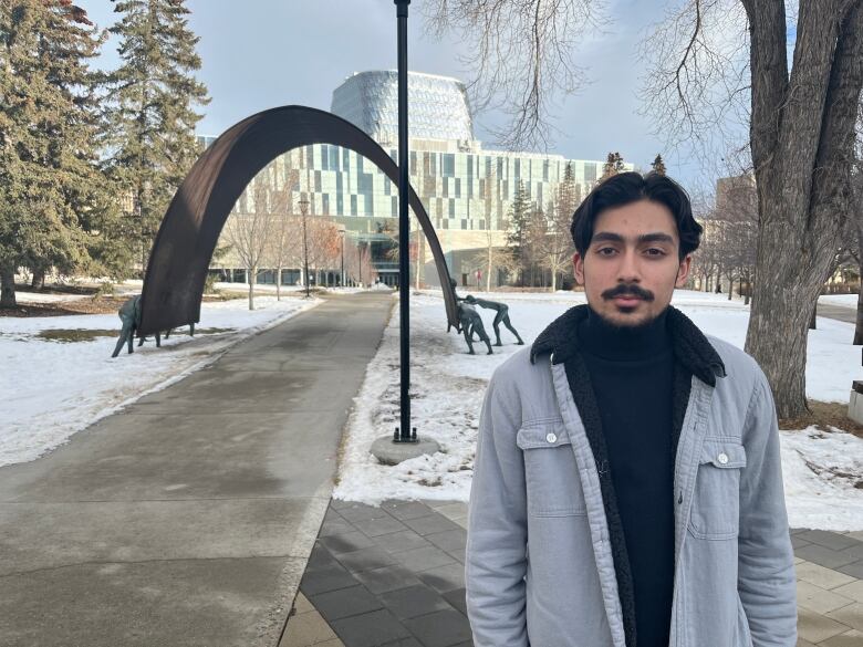 Shah stands outside in a grey jacket and dark blue shirt. His hands are in his pockets and a slight smile pulls at his lips as he looks into the camera. 