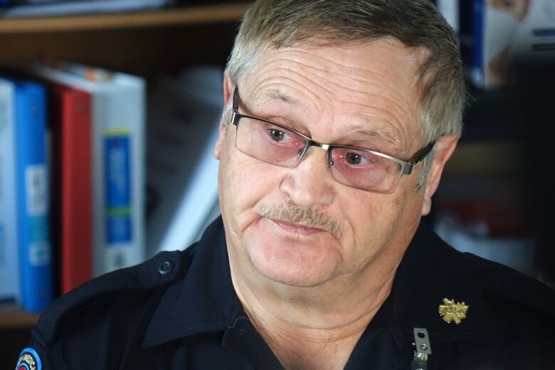 A man with short, greying hair, a moustache and glasses sits in front of a bookshelf full of binders.