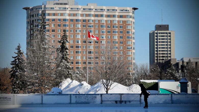 Someone skates on an outdoor rink with a Canadian flag and apartment building in the background.