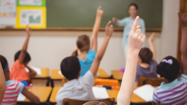 Blurred teacher teaching a lesson in class at the elementary school. Students have their hands up as you look at the back of their heads.