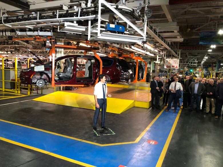 Trudeau answering questions at a microphone with a half built vehicle hoisted behind him