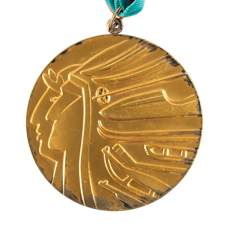 A gold medal, fraying at the edges, featuring the profiles of a laureated athlete and a First Nations indigenous person whose headdress is formed by winter sporting equipment. 