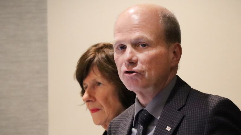 A man speaks as a woman behind him looks on.