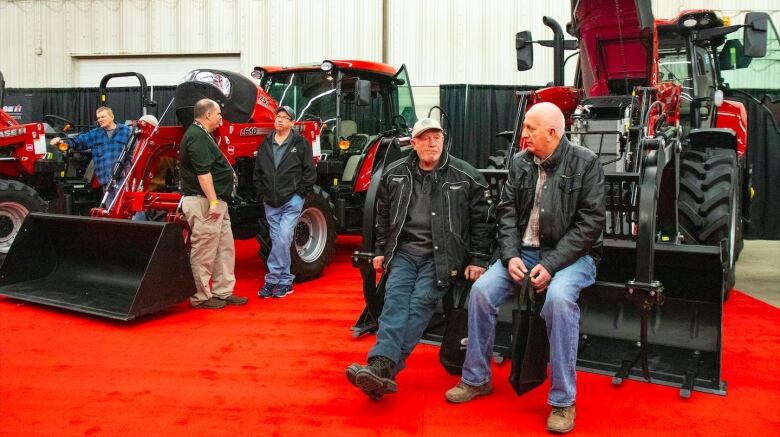 Two older men sit on the front loader of a tractor.