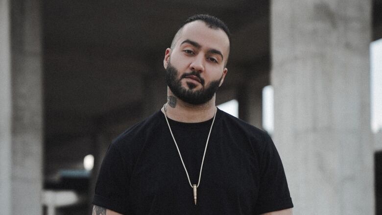 A man with dark hair and facial hair, wearing jeans, a black T-shirt and a silver necklace, poses for a portrait under an underpass.