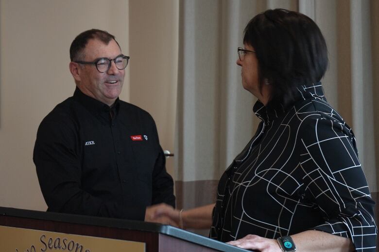 A man in a black shirt shaking hands with a woman in a black patterned shirt while standing at a podium. 