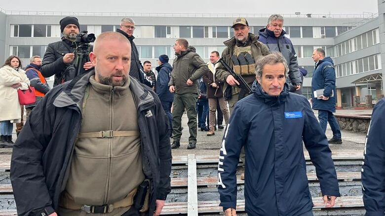 Two men in winter coats walk towards the camera, as journalists follow behind