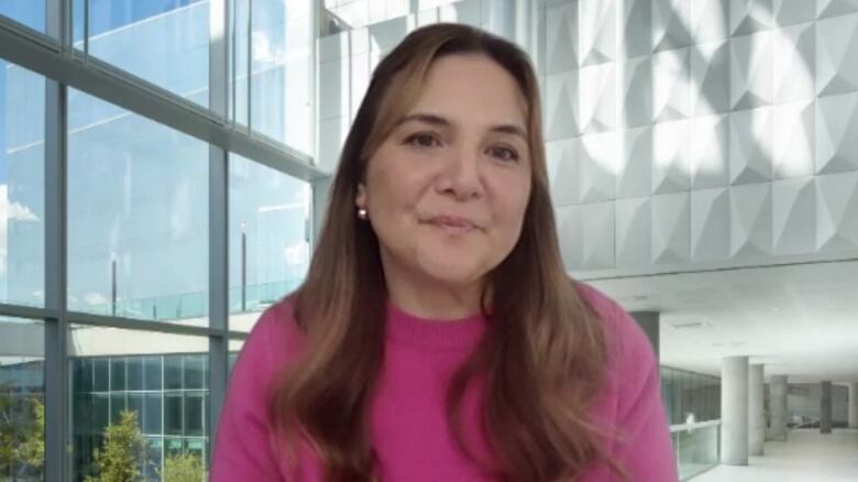 A woman with long brown hair and wearing a pink sweater is seen in front of a university background.