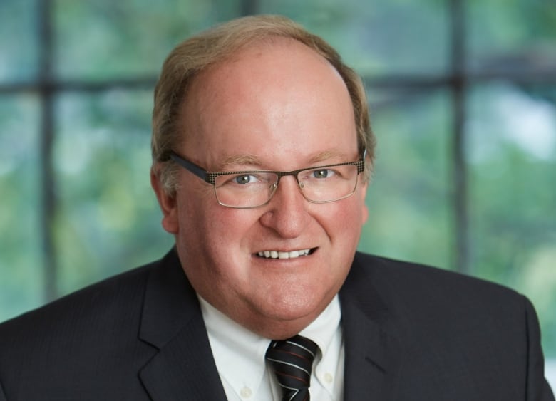 A bald spectacled man clad in a suit stands smiling.