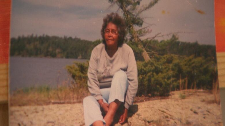 A woman in a gray sweater and light blue jeans sits on a rock by a lake.