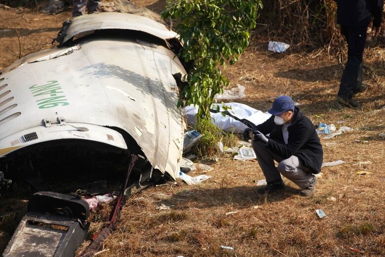 A man in a baseball cap and COVID mask crouches near a piece of fuselage from an airplane.