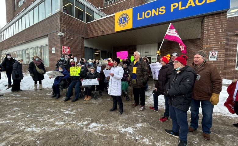 A crowd of people, some holding colourful signs, stand in front of a building with the name Lions Place in yellow letters on a blue background.