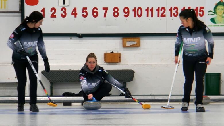 A woman slides on a curling sheet with a curling rock in her hand as two other curlers on her teams watch and carry brooms.