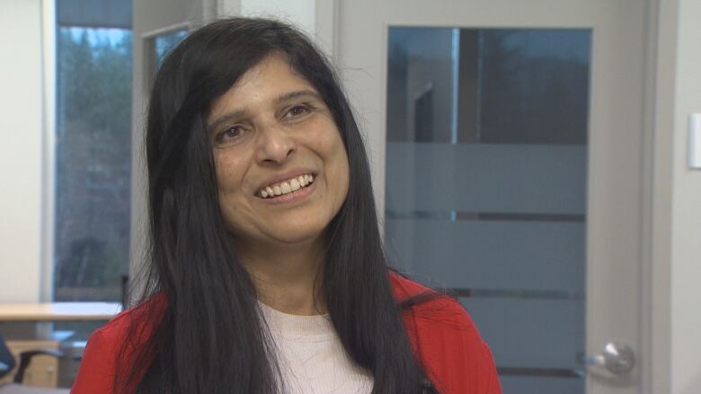 A woman stands in a red sweater with a window behind her as she talks about Nova Scotia Health's efforts to help nurses earn their credentials.