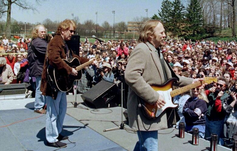 Three men are pictured from behind while playing guitar and singing to a crowd outdoors.