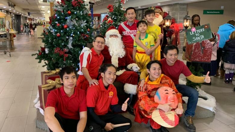 Eight men sit surrounding Santa Claus at a mall display with a Christmas tree in the background