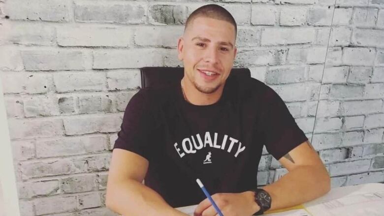 An Indigenous man sits with a form and black watch, with a black shirt that reads Equality. 