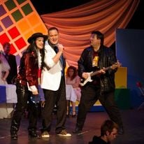 A man in a red shirt and black pants is at the front of a stage next to a man in a white coat and dark pants holding a microphone, with another man wearing a dark shirt, black leather jacket and dark pants playing a white electric guitar.