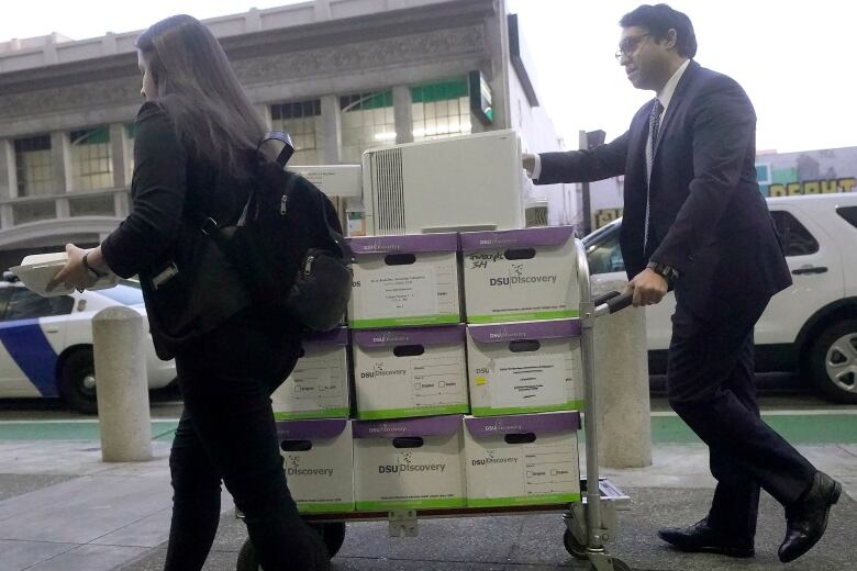 A woman, on the left, and a man, on the right, wheel a cart stacked with boxes along a sidewalk. 