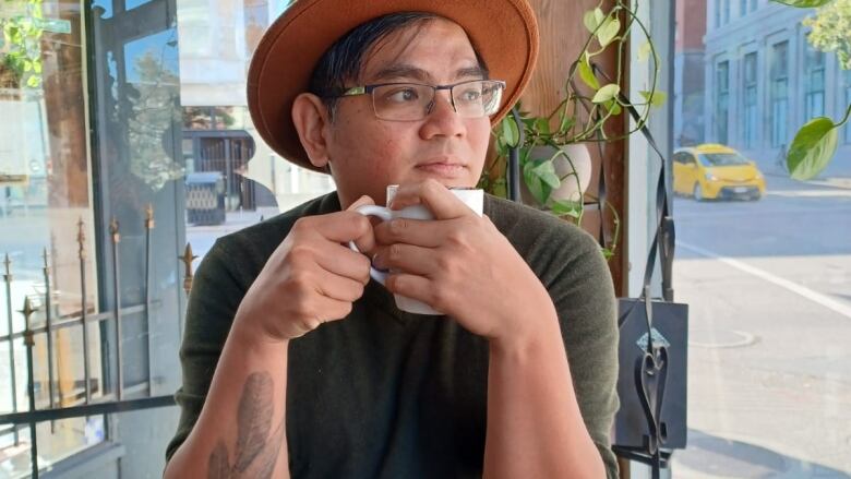 A man wearing a hat and holding a coffee cup stares into the distance.