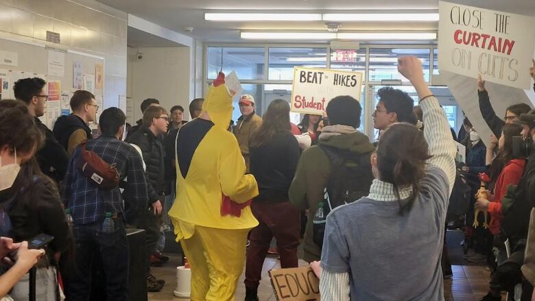 A gathering of students holding signs indoors. One man is wearing a chicken costume. 