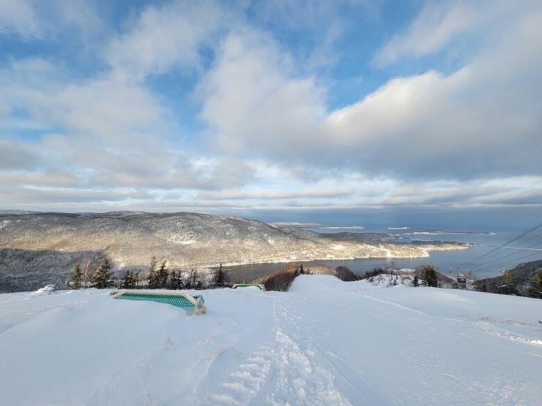 A snow-covered ski hill is shown with a view of the Atlantic ocean and blue clouds. 