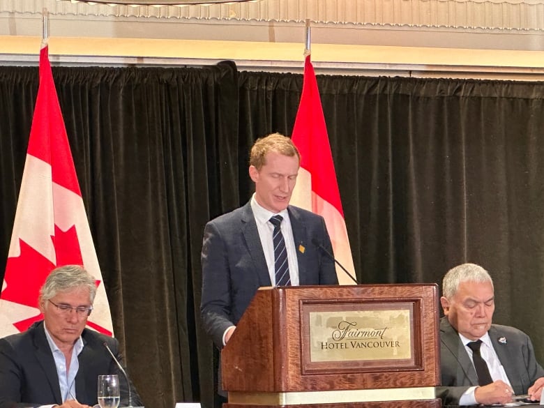 A white man speaks at a podium next to two men. Behind them, two Canada flags are draped.