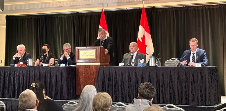 A man speaks at a podium surrounded by other people seated at tables. Two Canada flags are behind him.