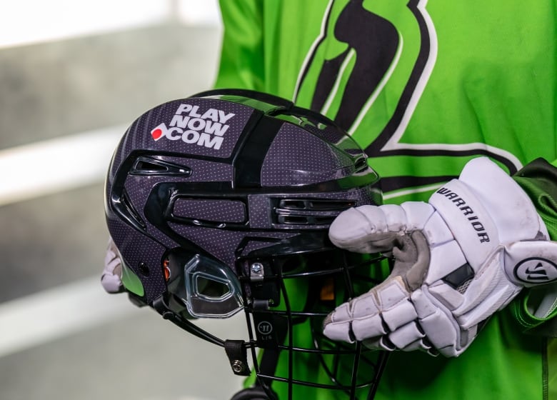 A lacrosse player with white gloves, and wearing a Saskatchewan Rush jersey, holds a black lacrosse helmet with the PlayNow.com logo on it.