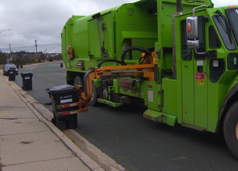 A garbage truck, on the right side of the frame, is parked on the street. A claw extended from the truck is about to close around a garbage container positioned against the curb. In the background, more garbage cans can be seen lined up against the curb.