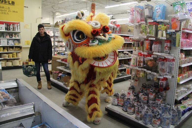 Two people in a stylized lion costume turn around the corner of an aisle at a Chinese supermarket, while a shopper carrying a handheld shopping cart, looks on. 