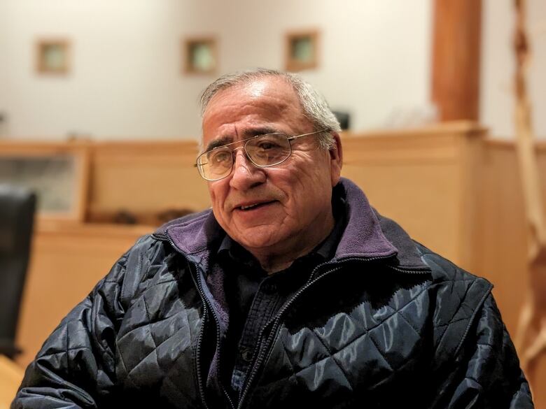 A man is sitting, wearing glasses and a puffy jacket. 