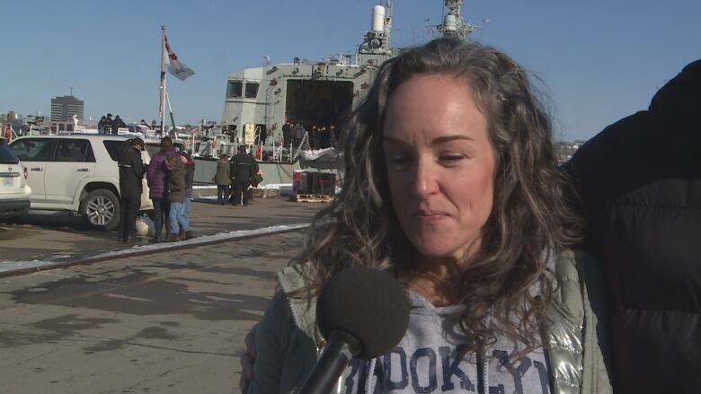 A tearful woman in a winter jacket looks down tearfully. There is a frigate in the background.