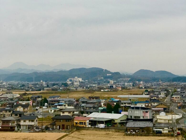 A scattering of low buildings are seen, with rounded mountains in the background and yellow fields in the foreground.