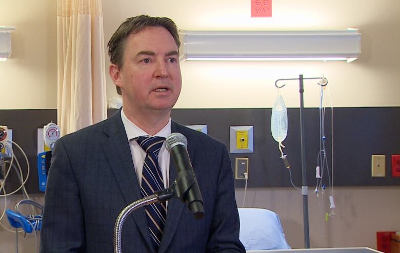 A man wearing a blue suit and blue striped tie speaks at a microphone set up in a hospital room. 