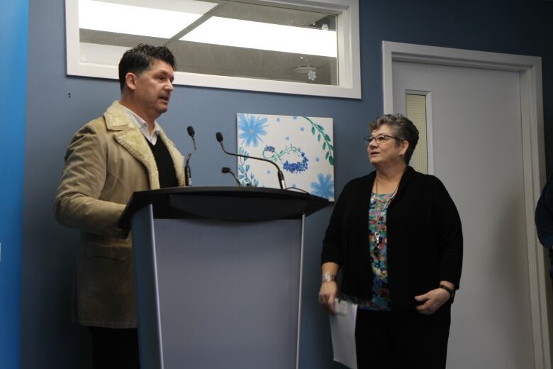 A man speaking at a podium with a woman standing beside him and looking at him.