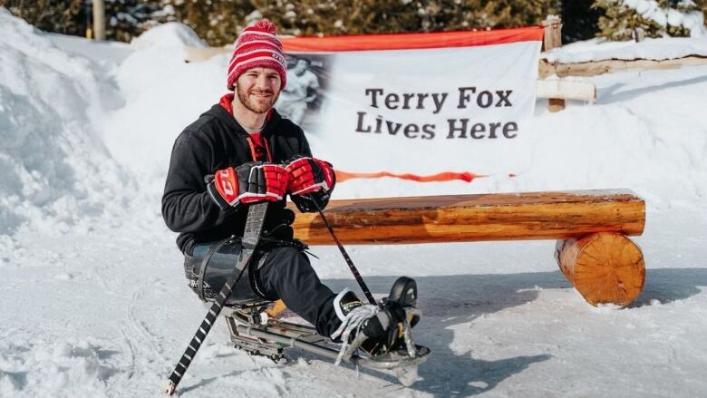 A man with a tuque and skating attire is pictured in the snow on a sledge in front of a banner that reads 'Terry Fox Lives Here.'